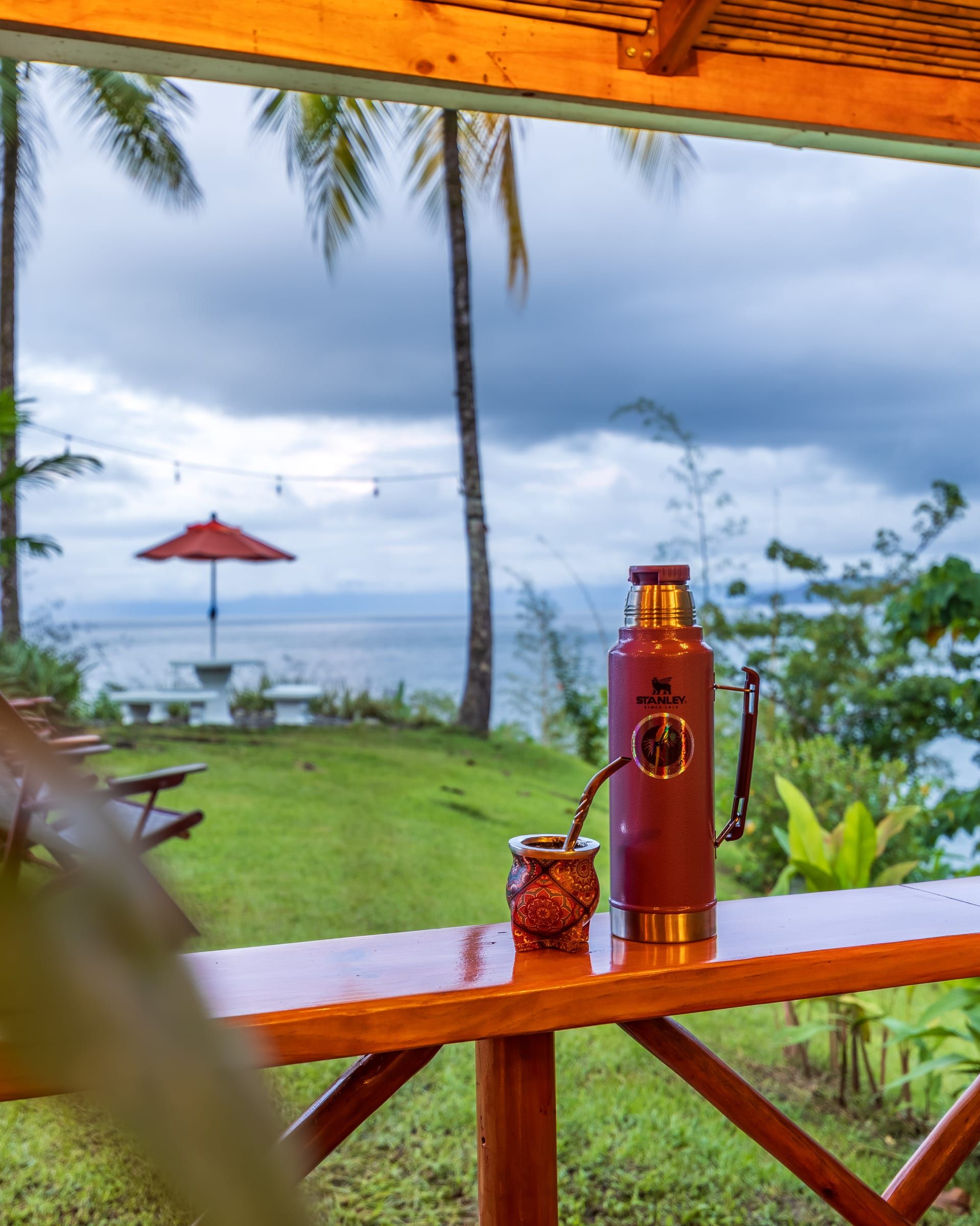 A thermos and a cup on a porch railing, with the garden and the sea beyond